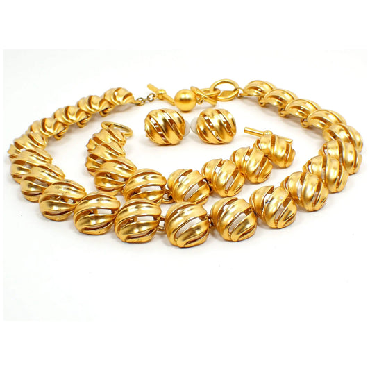 Angled front view of the retro vintage Anne Klein jewelry set. It is gold tone in color with domed oval links that have a ribbon like filigree cut out design that goes diagonally across each link. There is a necklace and a bracelet that have toggle clasps and a pair of clip on earrings. 