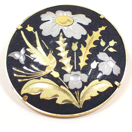 Enlarged view of the retro vintage round Damascene brooch pin. There are metallic silver, brass, and gold color flowers and leaves with a gold color bird engraved on a black background. 