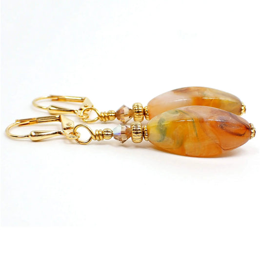 Side view of the handmade lucite drop earrings made with vintage beads. The metal color is gold plated. There are new faceted glass crystal beads at the top in a light orange color. The bottom lucite beads are long faceted oval shape and have marbled colors that are mostly peach, orange, yellow, and brown, but have some other colors swirled in as well. Each bottom bead is different in colors and pattern than the other.