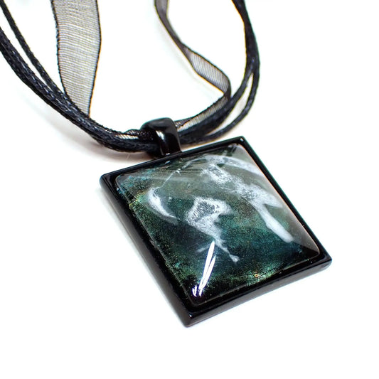 Enlarged view of the square handmade frost resin pendant. The necklace part has three strands of black faux leather cord and a strand of black organza ribbon. There is a black coated square pendant at the bottom with a domed resin cab. The resin has iridescent shades of green and yellow and an abstract frost design.