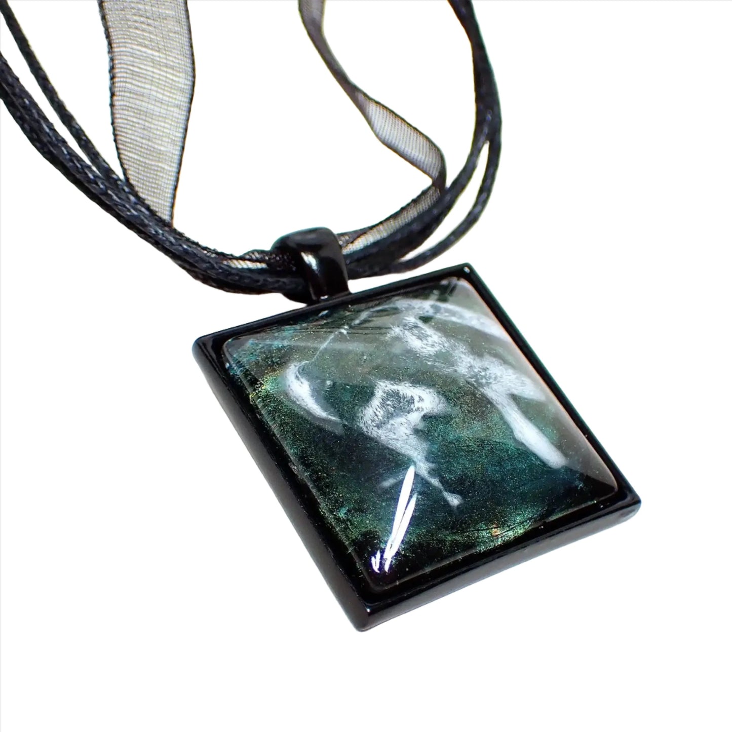 Enlarged view of the square handmade frost resin pendant. The necklace part has three strands of black faux leather cord and a strand of black organza ribbon. There is a black coated square pendant at the bottom with a domed resin cab. The resin has iridescent shades of green and yellow and an abstract frost design.
