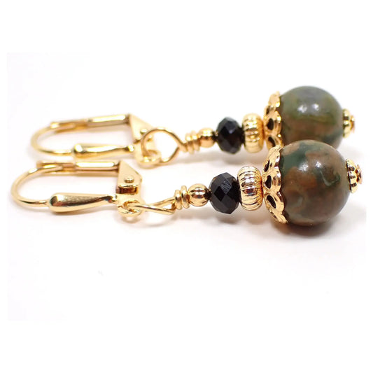 Enlarged view of the handmade rainforest jasper earrings. The metal is gold plated in color. There are small faceted black crystal bicone beads at the top. The bottom gemstone beads are round sphere shaped and have marbled shades of green and brown.