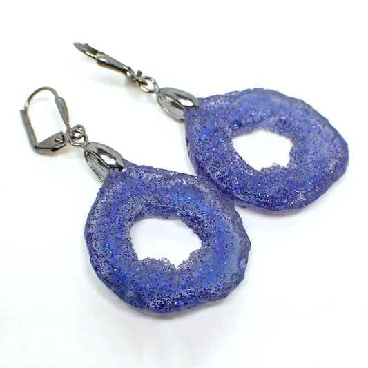 Angled front view of the handmade druzy geode style earrings. The metal is gunmetal gray in color. The drops are rounded teardrop shape with a druzy like freeform edge. The resin has blue glitter throughout.