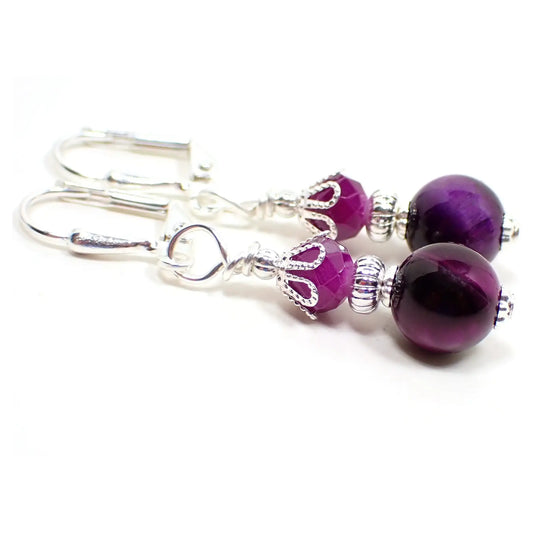 Angled view of the handmade tiger's eye gemstone earrings. The metal is silver plated in color. There are faceted glass purple beads at the top. The bottom tiger's eye beads are small round ball shaped and are dyed a dark purple in color.