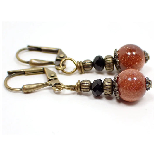 Side view of the handmade goldstone drop earrings. They have antiqued brass color beads and findings. There are faceted black crystal glass bicone beads at the top and small round ball goldstone beads at the bottom. The goldstone beads are orange glass with tiny flecks of copper for sparkle.