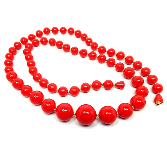 Angled view of the Mid Century vintage beaded necklace. The beads are bright red in color. There are small gold tone plated beads in between them. All of the beads are round with the largest at the bottom and going smaller as the necklace goes up. The clasp is a screw barrel clasp with bright red lucite around it.