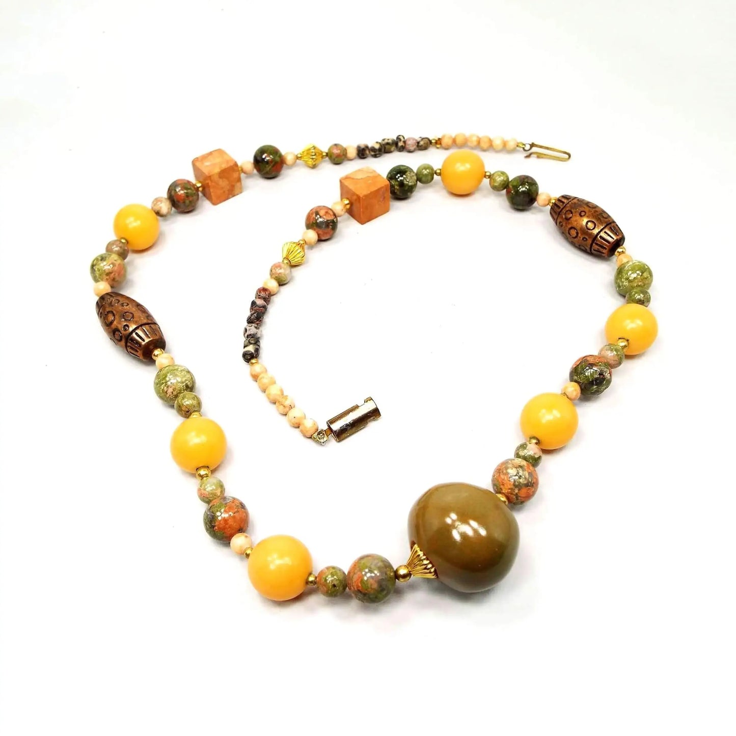 Angled view of the asymmetrical chunky beaded retro vintage Suzette necklace. There is a slide in style clasp at the end. The metal beads are gold tone and copper tone in color. There are oval, square, bicone, and round shaped beads. The colors vary with mostly green, orange, pin, and brown tones. There are porcelain, plastic, metal, and gemstone beads.