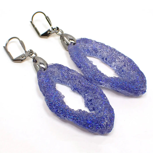 Angled front view of the handmade oval shaped faux geode druzy slice style earrings. The metal is dark gunmetal gray in color. The resin has sparkly blue glitter throughout.