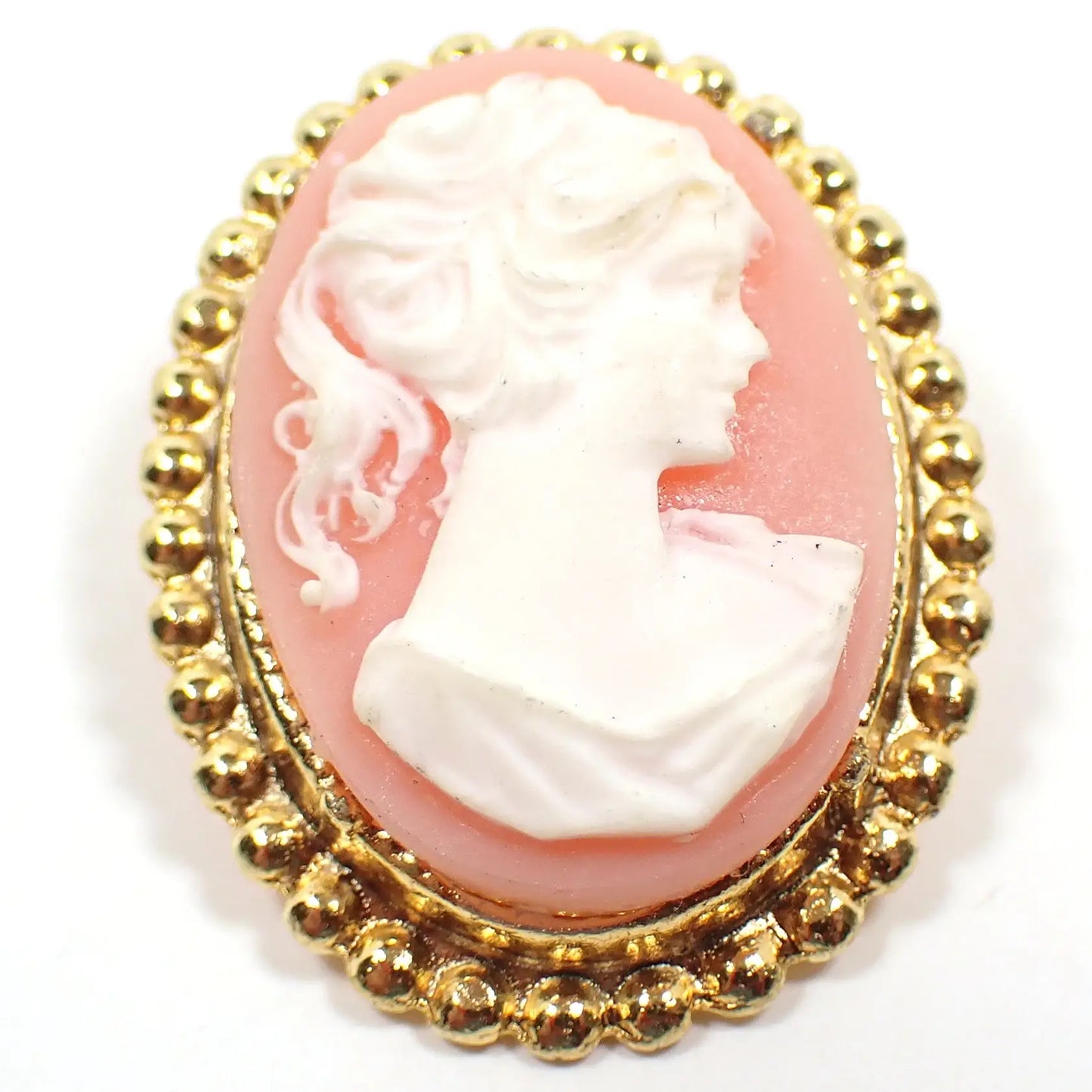 Enlarged front view of the retro vintage girl cameo brooch pin. The metal is gold tone in color. It is oval shaped with a dotted edge. There is a bust of a girl with a pony tail on the front with a light pink background.