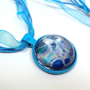 Enlarged view of the handmade resin pendant necklace. The necklace part has a strand of blue organza, which is like a wider mesh ribbon. There are also three strands of waxed cord in aqua blue. The pendant is round and aqua blue enameled. There is a resin cab on the front that has an abstract design with shades of blue and iridescent areas. A bubble on the edge can be seen on the left hand side.