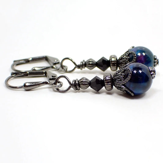 Side view of the small handmade dyed tiger's eye earrings. The metal is gunmetal gray in color. There are small faceted black crystal glass beads at the top. The gemstone beads are round sphere shaped and are dyed blue with small areas of purple.