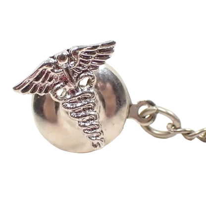 Enlarged angled front view of the retro vintage Caduceus tie tack. It is silver tone in color and is the symbol for medical professionals with two snakes wrapped around a staff with wings at the top.