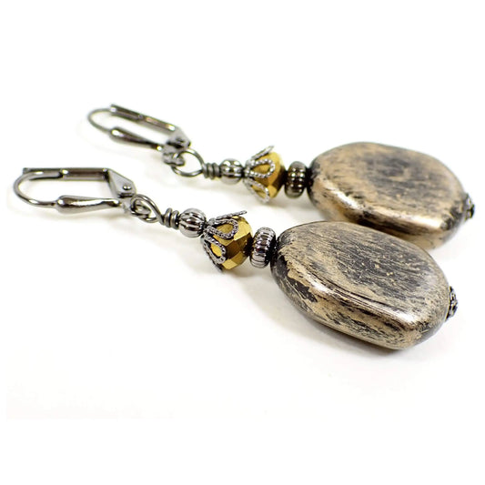 Angled view of the handmade drop earrings. The metal is gunmetal gray in color. There are faceted glass beads at the top in a dark antiqued metallic gold color. The bottom beads are flat irregular shaped similar to a teardrop with a faceted edge. The acrylic beads are black with an antiqued metallic gold paint brushed on them.