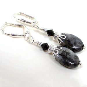 Angled view of the handmade gemstone earrings. The metal is silver plated in color. There are black faceted glass crystal beads at the top. The bottom beads are larvikite, which is also sometimes called blue labradorite. They are puffy oval shaped and are primarily gray with marbled areas of black. There are a few areas on each bead that has flashes of silvery or blue color as you move around in the light.