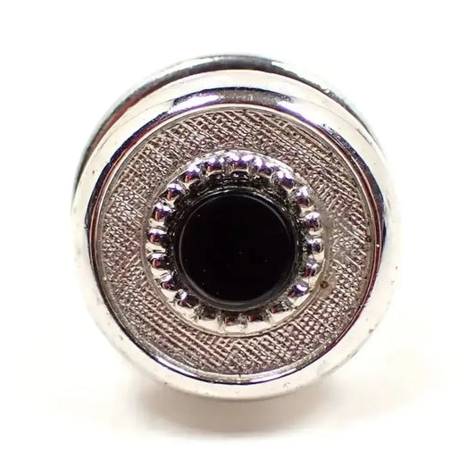 Front view of the Mid Century vintage tie tack. It is round and silver tone plated in color. The front has a small round black plastic cab surrounded by a textured matte silver color edge. The back clutch is the kind without a chain on it.