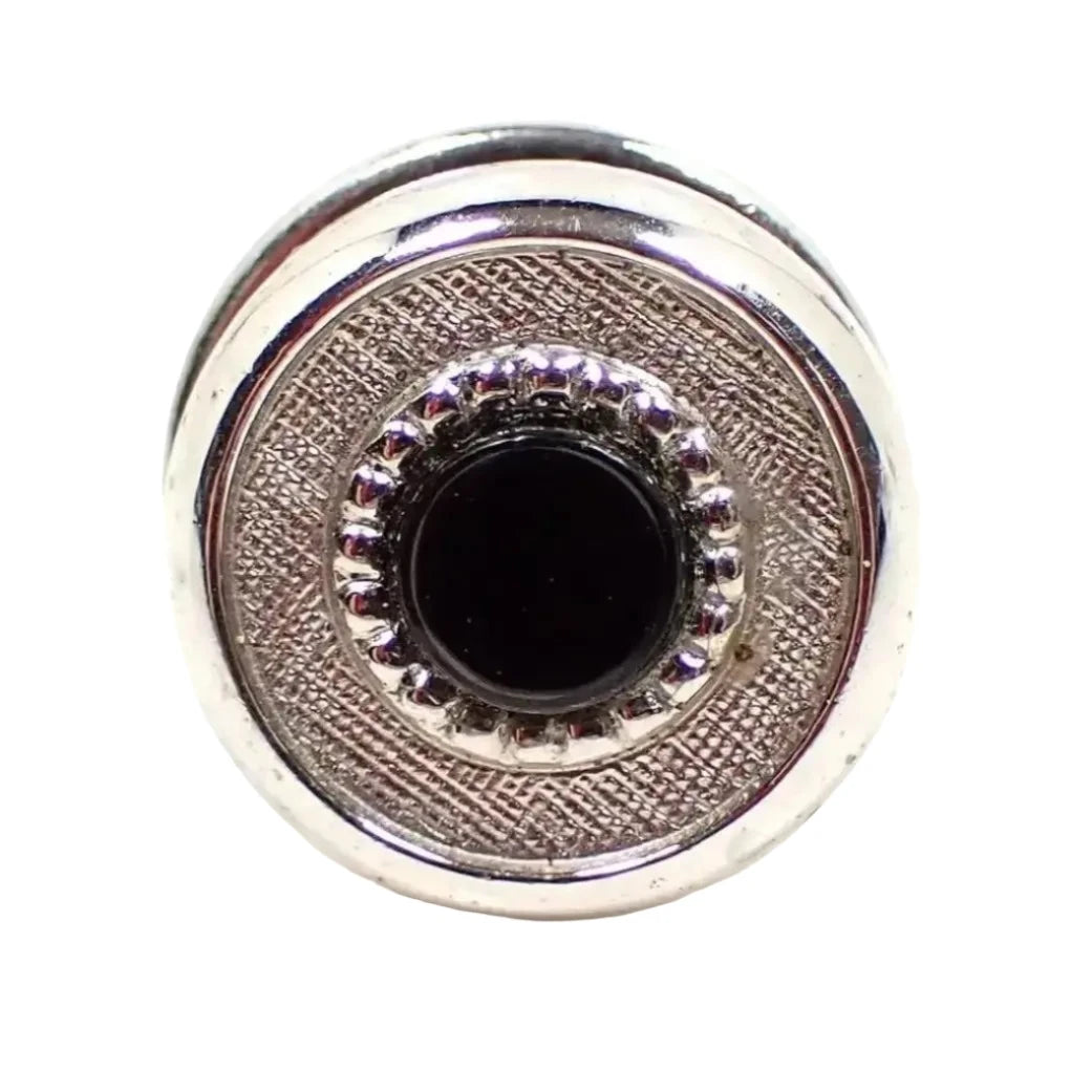 Front view of the Mid Century vintage tie tack. It is round and silver tone plated in color. The front has a small round black plastic cab surrounded by a textured matte silver color edge. The back clutch is the kind without a chain on it.