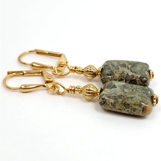 Angled view of the handmade rhyolite gemstone earrings. The metal is gold plated in color. There are two small metal beads at the top of the earrings. The bottom bead is rhyolite gemstone and is a puffy rectangle shape with rounded corners. There are blocks and marbled areas with different shades of mossy green and some brown tones.