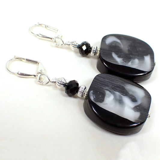 Angled view of the handmade earrings with vintage Italian lucite beads. The metal is silver plated in color. There are black faceted glass crystal beads at the top. The bottom lucite beads are a slice style with rounded edges. There is black lucite on each side edge of the beads and the middle has translucent area with swirls of black and white with hints of gray.