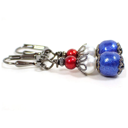 Enlarged side view of the red white and blue handmade earrings. The metal is gunmetal gray in color. There is a small metallic red glass bead at the top, a white faux pearl bead in the middle, and a sparkly blue bead at the bottom.