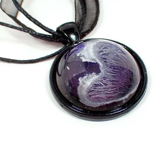 Enlarged view of the handmade resin pendant. The necklace part shows three black strands of faux leather cord and a strand of organza ribbon. There is a black coated round pendant with a domed resin cab. The resin has an iridescent purple background and an abstract frost design in white through it.