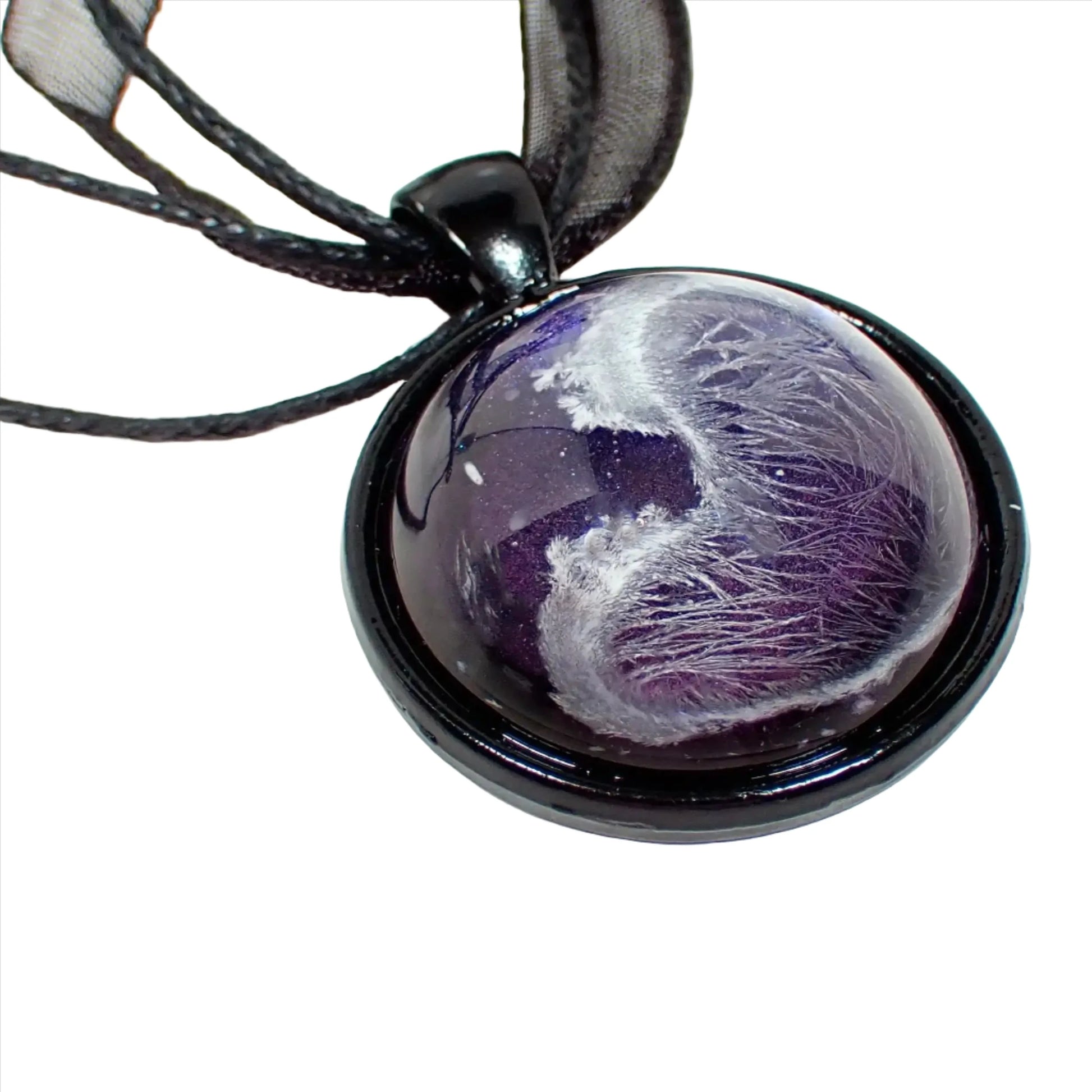 Enlarged view of the handmade resin pendant. The necklace part shows three black strands of faux leather cord and a strand of organza ribbon. There is a black coated round pendant with a domed resin cab. The resin has an iridescent purple background and an abstract frost design in white through it.