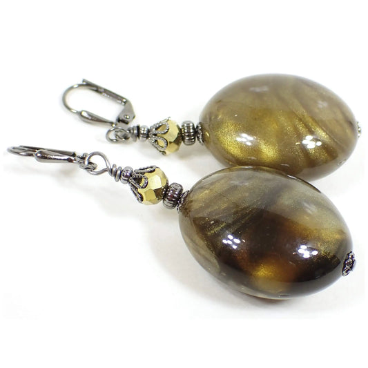 Angled view of the big oval handmade drop earrings with vintage lucite beads. The metal is gunmetal gray in color. There are faceted metallic bronze glass beads at the top. The bottom beads are large puffy ovals that have marbled swirls in shades of pearly brown and olive green.