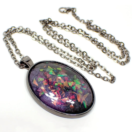 Enlarged front view of the large oval handmade resin pendant. The metal is gunmetal plated in color. The large oval pendant has a resin cab with marbled pearly shades of dark gray and purple. There are chunky pieces of iridescent glitter embedded in the resin that flash and sparkle with different colors depending on how the light hits it.