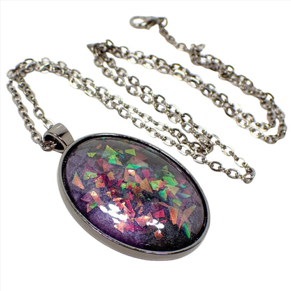 Enlarged front view of the large oval handmade resin pendant. The metal is gunmetal plated in color. The large oval pendant has a resin cab with marbled pearly shades of dark gray and purple. There are chunky pieces of iridescent glitter embedded in the resin that flash and sparkle with different colors depending on how the light hits it.