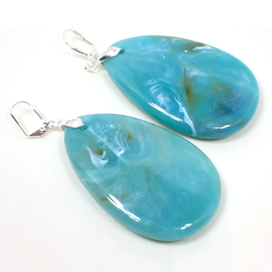 Angled view of the big heavy acrylic teardrop earrings. The metal is silver plated in color. There are large teardrop shaped drops that are aqua blue in color with small marbled swirls of white and brown.