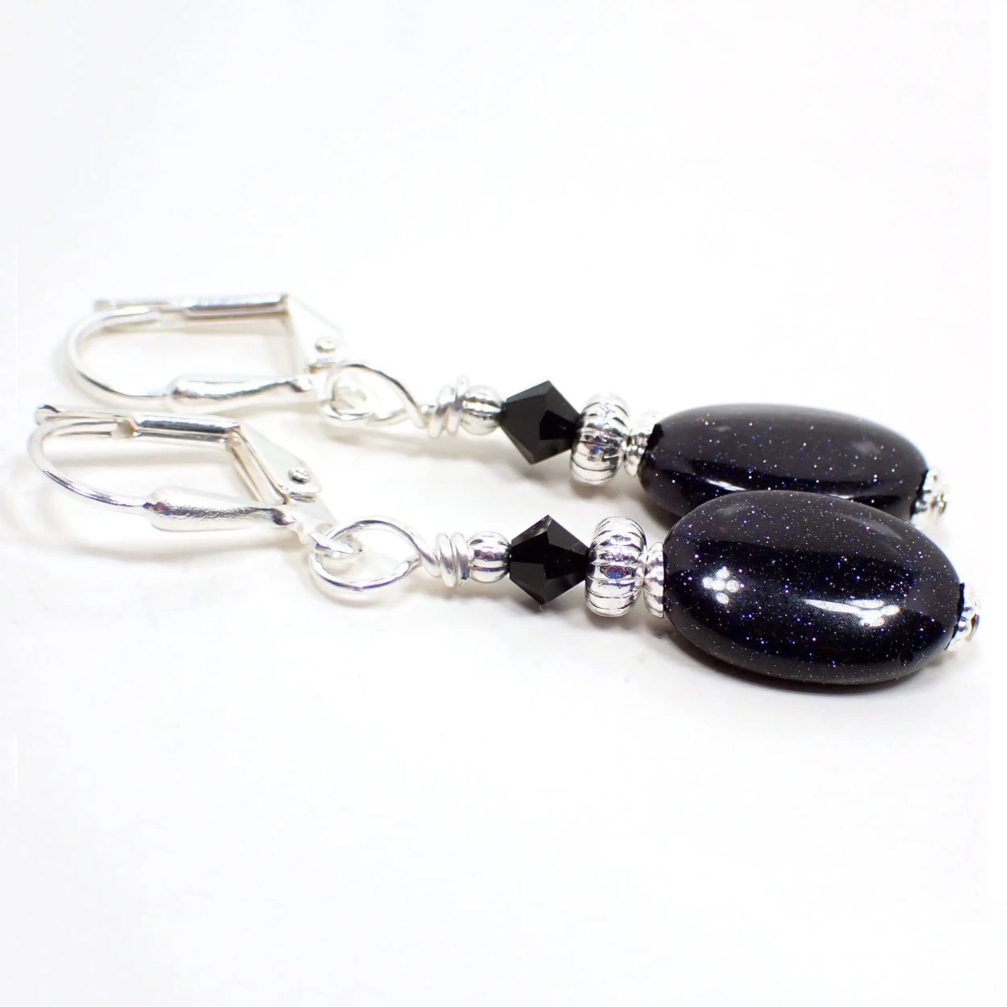 Angled view of the handmade blue goldstone drop earrings. The metal is silver plated in color. There are small black glass crystal beads at the top. The bottom beads are puffy oval in dark blue with tiny flecks of metallic blue and silver glitter in the glass.
