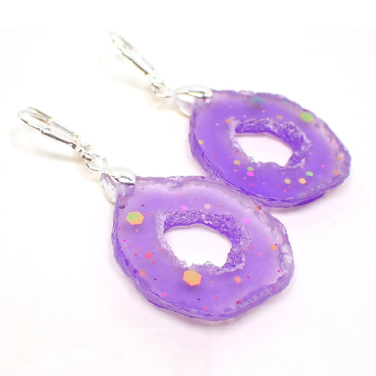 Angled front view of the handmade geode slice style earrings. The metal is silver tone plated in color. The drops are rounded teardrop shape with druzy like edges. They are a bright almost semi translucent pearly purple in color. The resin has specks of chunky iridescent glitter.