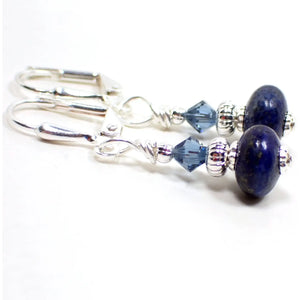 Angled view of the small handmade lapis lazuli drop earrings. The metal is silver plated in color. There are blue smoky blue faceted glass crystal beads at the top. The bottom gemstone beads are a dark blue in color with tiny flecks of gold color. The beads are rondelle saucer like shaped.