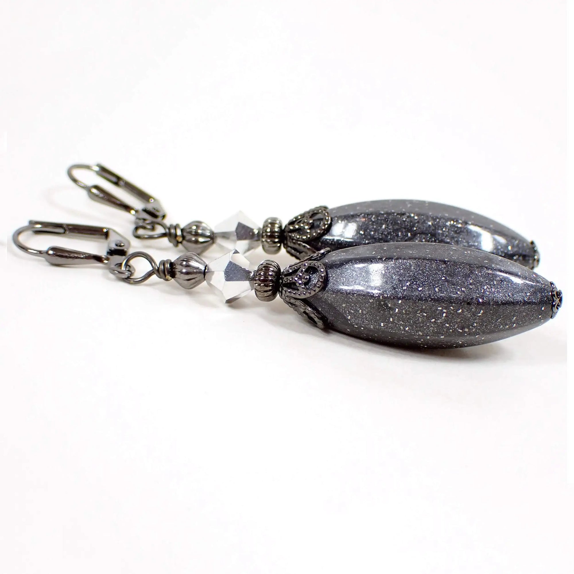 Angled view of the large handmade drop earrings with vintage lucite beads. The metal is gunmetal gray in color. There are faceted clear and metallic silver glass crystal beads at the top. The bottom lucite beads are a long faceted oval shape and are pearly dark gray in color with tiny flecks of glitter embedded in them.