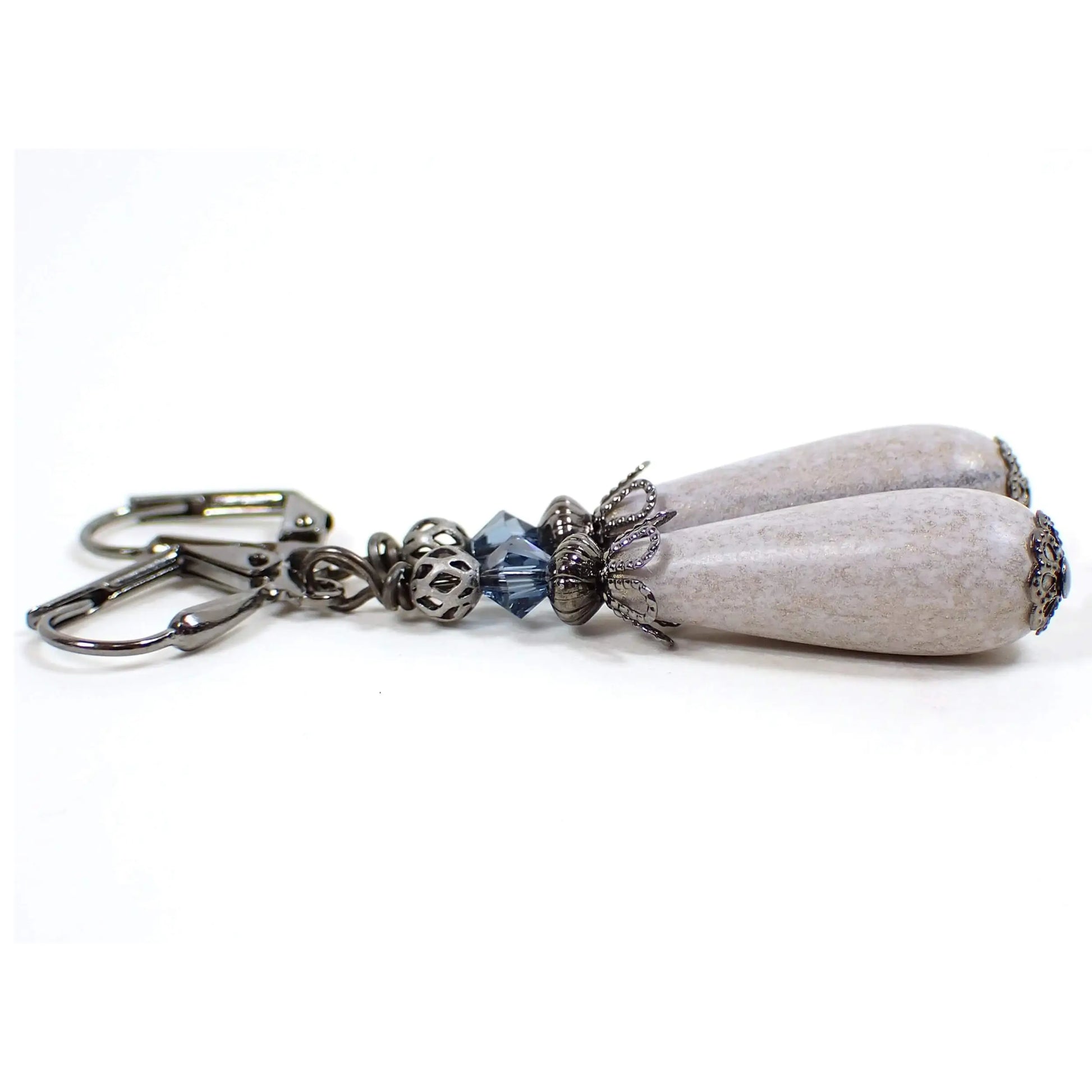 Side view of the handmade earrings with vintage lucite beads. The metal is gunmetal gray in color. There are dark blue faceted glass crystal beads at the top. The bottom vintage lucite beads are teardrop shaped and are a light gray color with light golden sheen on the outside.