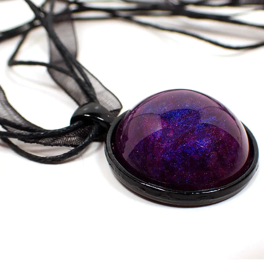 Enlarged view of the handmade goth pendant necklace. The necklace part is a strand of black organza which is a semi transparent ribbon like design, and three strands of waxed cord. The pendant is round and black enameled. There is a handmade domed resin cab on the front that is mostly dark pearly purple with some areas of dark blue and pink as well.