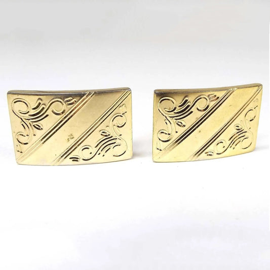 Front view of the Mid Century vintage Anson etched cufflinks. The metal is gold tone in color and they are a slightly curved rectangle shape. There is an etched design of diagonal lines and floral type curls on the front.