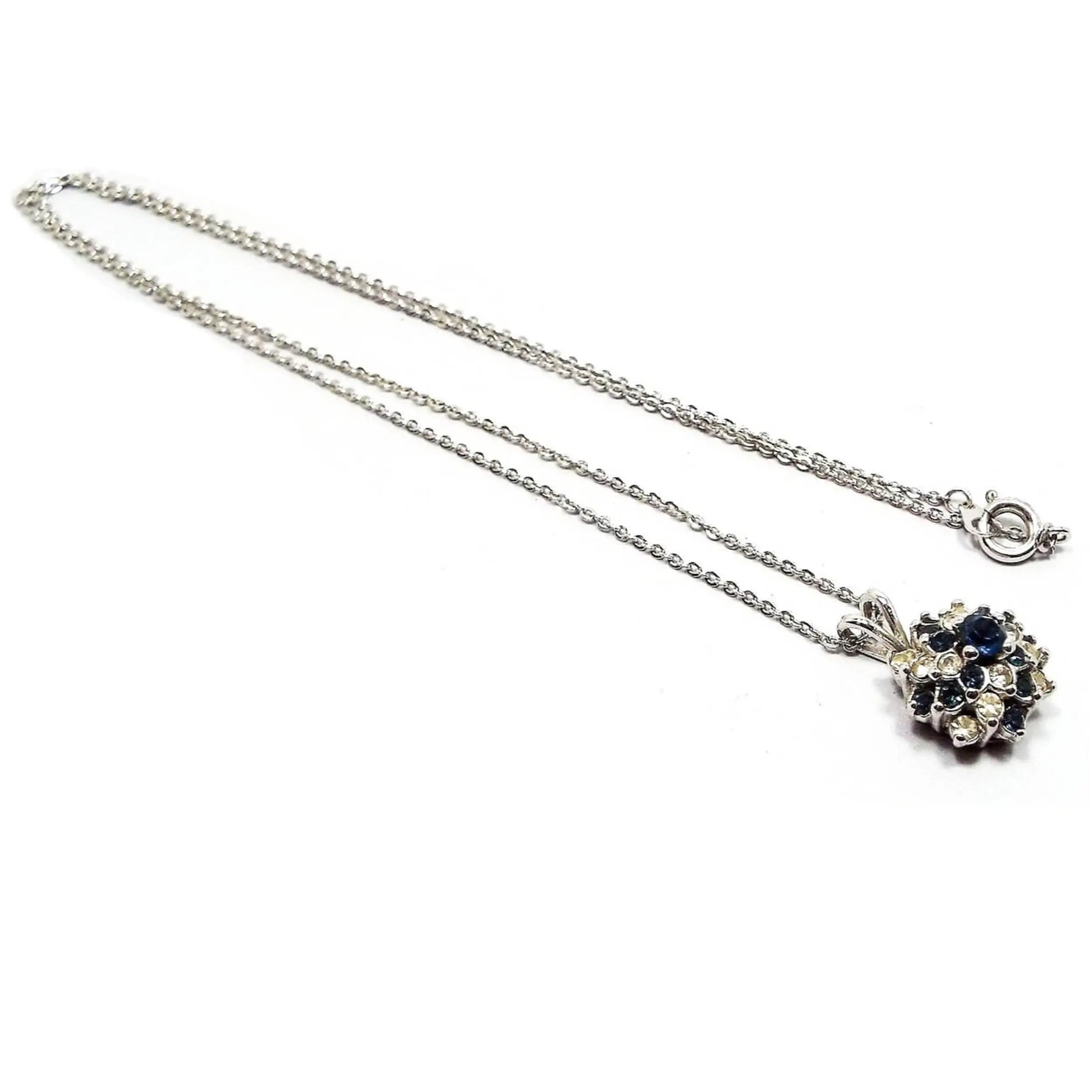Front view of the small retro vintage rhinestone pendant necklace. The metal is silver tone in color and it has a thinner sized cable chain with a spring ring clasp at the end. The bottom pendant is round flower like stacked shape with dark blue and clear rhinestones.