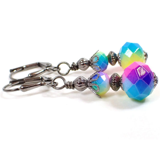 Side view of the rainbow earrings. The metal is gunmetal gray in color. There are faceted glass rondelle beads at the top and larger ones at the bottom. They have different vibrant colors of the rainbow blended all the way around the beads.