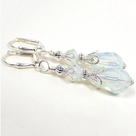 Angled view of the handmade teardrop earrings. The metal is silver plated in color. The glass crystal beads are a semi translucent opalescent kind of white in color. There is a faceted rondelle bead at the top and a teardrop shaped bead at the bottom.