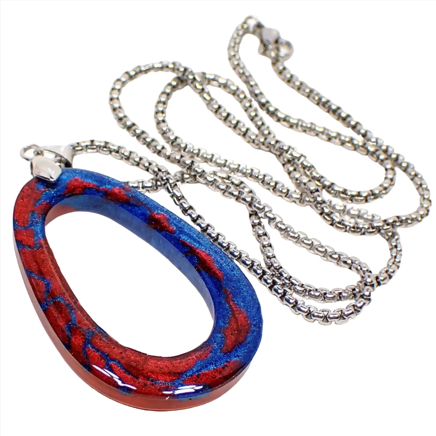Angled enlarged view of the handmade resin pendant necklace. The metal is antiqued silver tone in color. The chain is box style with a lobster claw clasp. The resin pendant is an open oval shape with pearly metallic blue and red resin. Each side side of the front of the pendant has one of the main colors with small splashes of the other color throughout it.