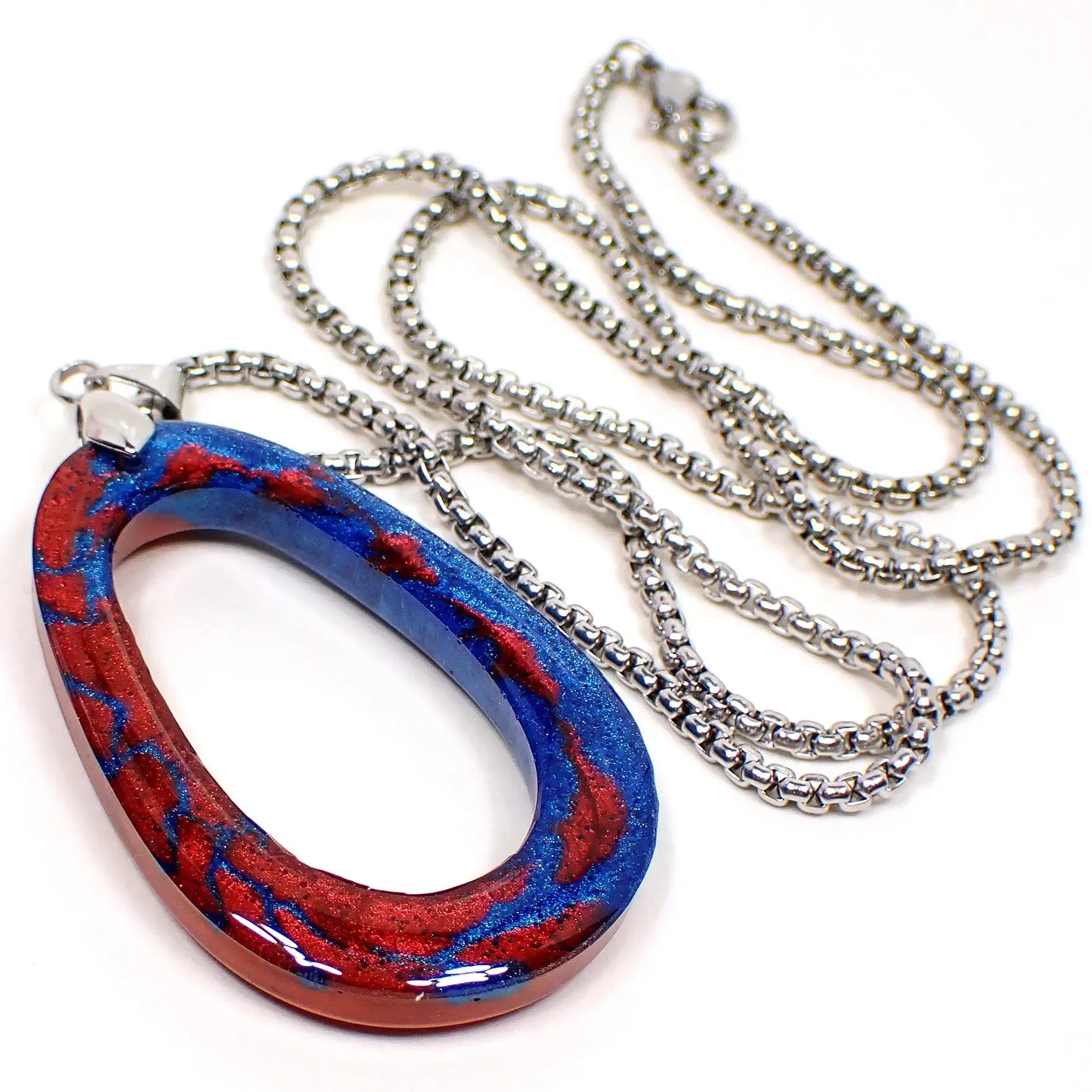 Angled enlarged view of the handmade resin pendant necklace. The metal is antiqued silver tone in color. The chain is box style with a lobster claw clasp. The resin pendant is an open oval shape with pearly metallic blue and red resin. Each side side of the front of the pendant has one of the main colors with small splashes of the other color throughout it.