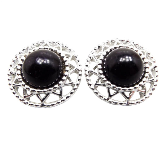 Front view of the retro vintage Sarah Coventry clip on earrings. The metal is silver tone in color. they have a round filigree sunlike design with a domed black plastic cab in the middle.