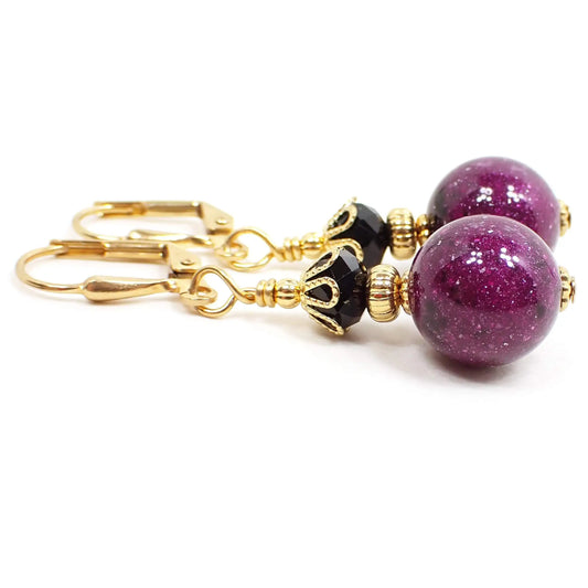 Side view of the handmade galaxy drop earrings. The metal is gold plated in color. The bottom round beads are sparkly purple with specks of black here and there. There are black faceted glass beads at the top.