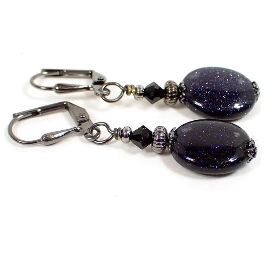 Side view of the handmade goldstone earrings. The metal is gunmetal gray in color. There are black faceted glass beads at the top. The bottom goldstone beads are dark blue puffy oval shaped with tiny flecks of metal glitter in the glass.