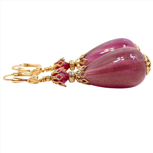 Side view of the large teardrop handmade earrings with vintage lucite beads. The metal is gold plated in color. There are faceted glass crystal beads at the top in a bright fuchsia purple color. The bottom lucite teardrop beads are purple with a metallic gold sheen.