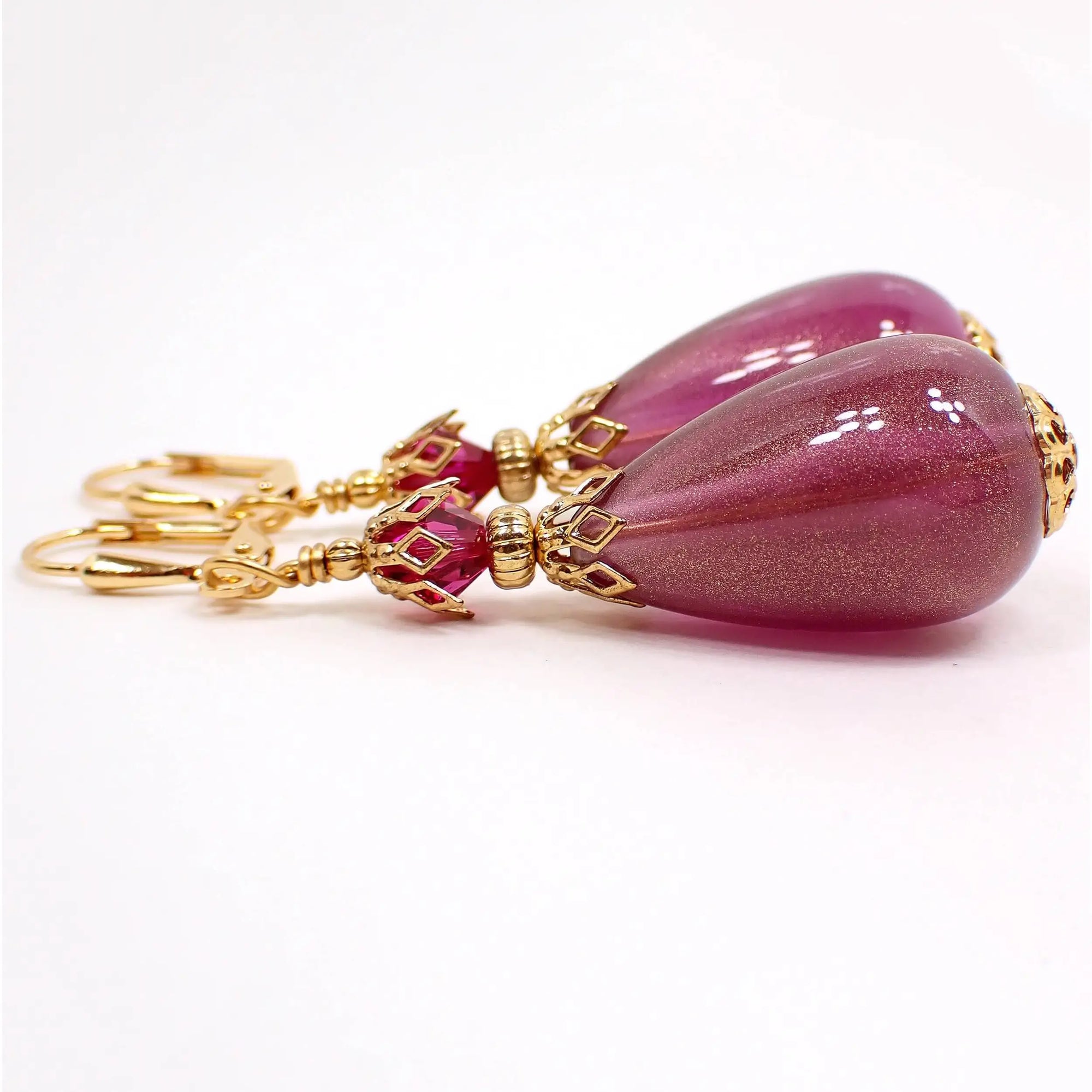 Side view of the large teardrop handmade earrings with vintage lucite beads. The metal is gold plated in color. There are faceted glass crystal beads at the top in a bright fuchsia purple color. The bottom lucite teardrop beads are purple with a metallic gold sheen.
