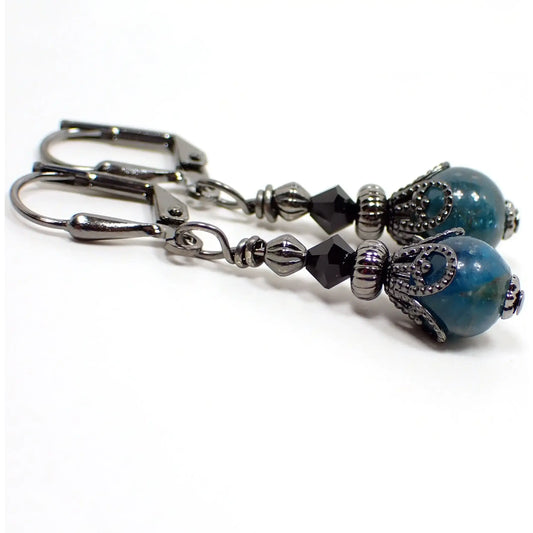 Photo of the small handmade apatite drop earrings. The metal is gunmetal gray in color. There is a faceted glass crystal bead at the top and a small round ball apatite gemstone bead at the bottom. The gemstone is a marbled teal blue in color.