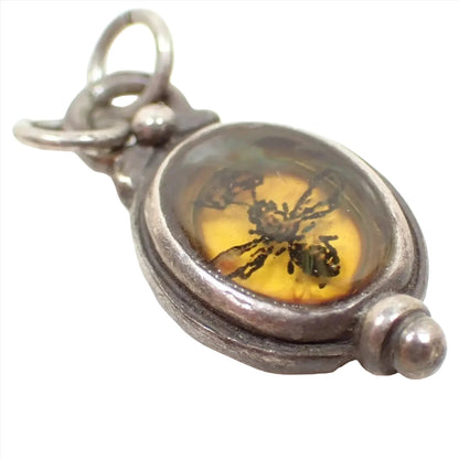 Enlarged angled view of the pre owned modern Queen Bee charm. The metal is antiqued silver in color. The sterling setting is oval shaped with a round ball part on the bottom and a loop on the top. There is yellow resin in the middle with semi translucent swirls at the top. There is a printed decal of a bee with a crown embedded inside.