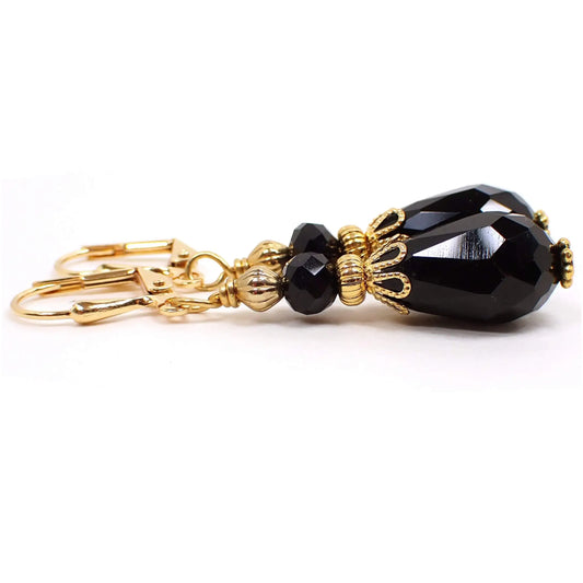 Angled side view of the handmade vintage style black teardrop earrings. The metal is gold plated in color. There is a faceted glass rondelle black bead at the top and a faceted teardrop black bead at the bottom.