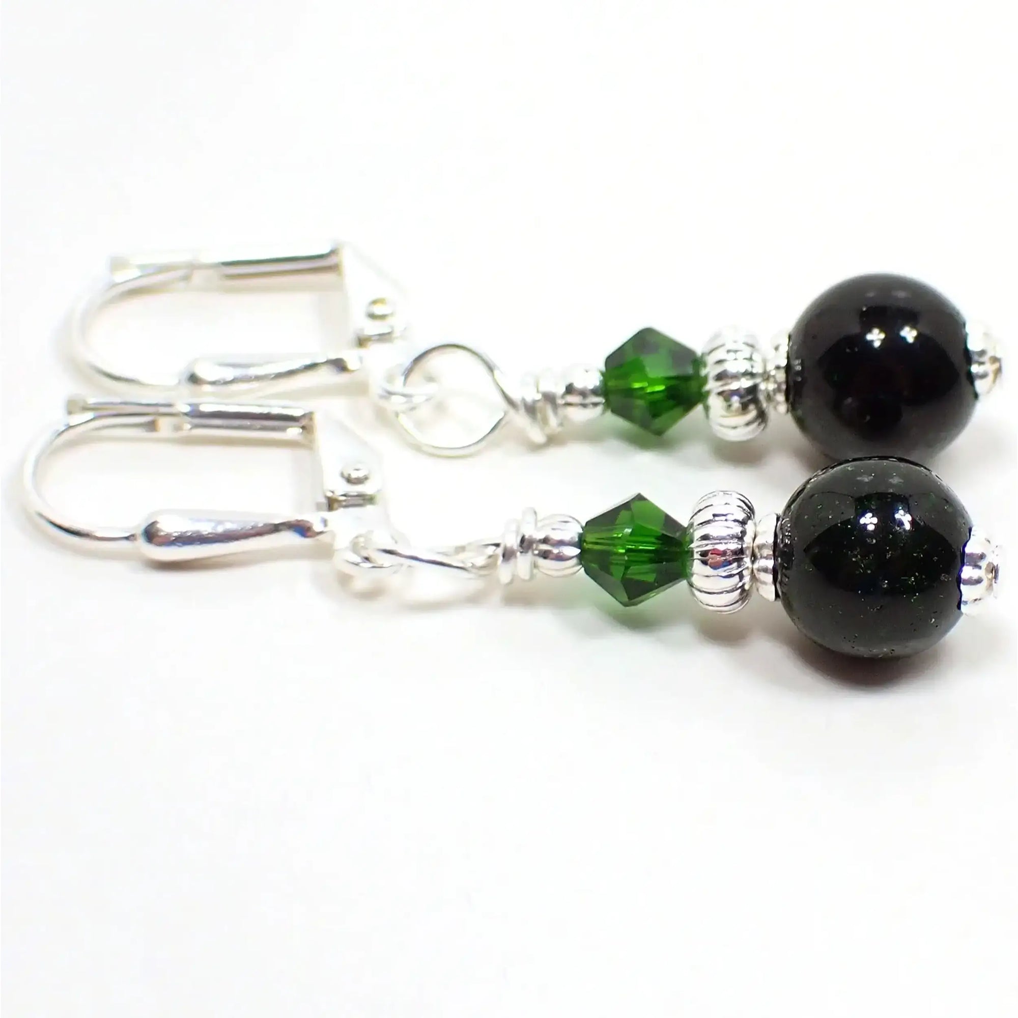 Side view of the small handmade green goldstone earrings. The metal is silver plated in color. There are small faceted green glass crystal beads at the top. The bottom beads are round sphere shaped and are dark green in color with tiny flecks of metallic green for sparkle.